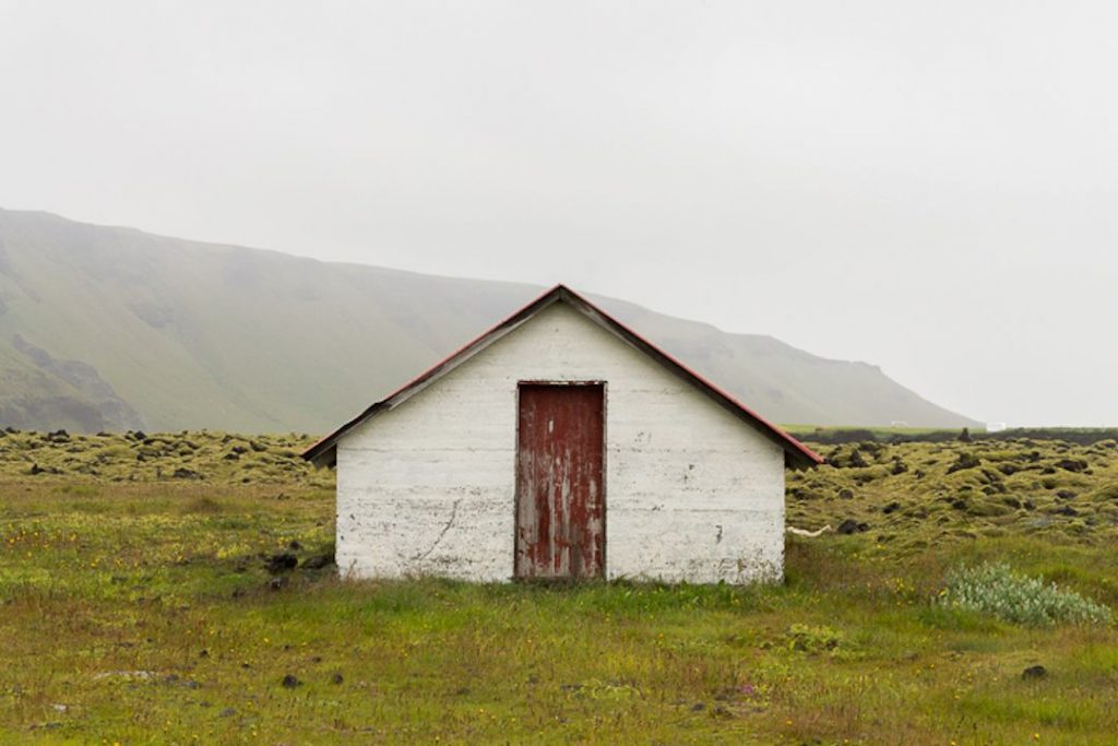 small white shack with a rusting red door in a green pasture