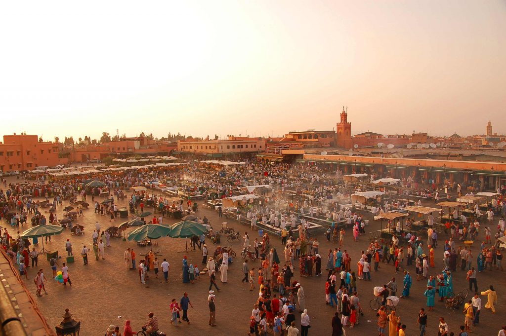 a colourful crowded market square with a romantic sunset looming in the background 