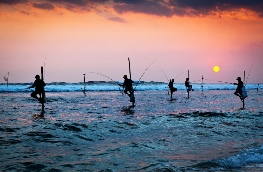 Several stilt fishermen are perched in the sea below and orange-hued sky