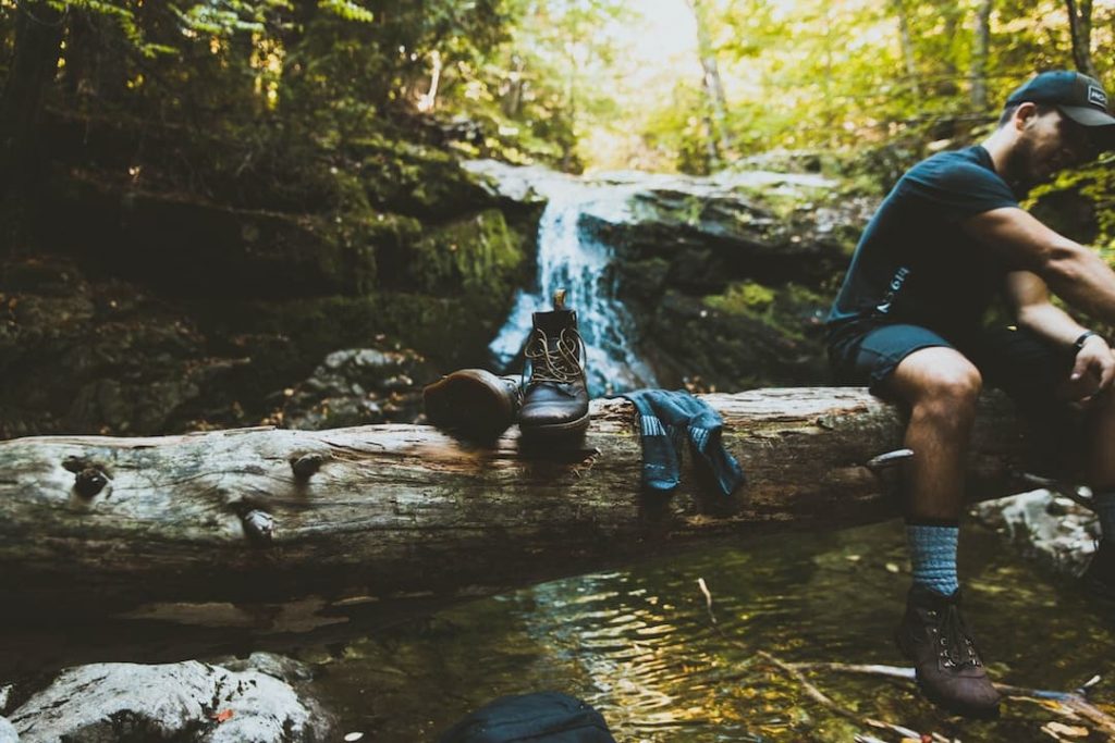 A man sitting on a log over running water, with a pair of hiking boots beside him