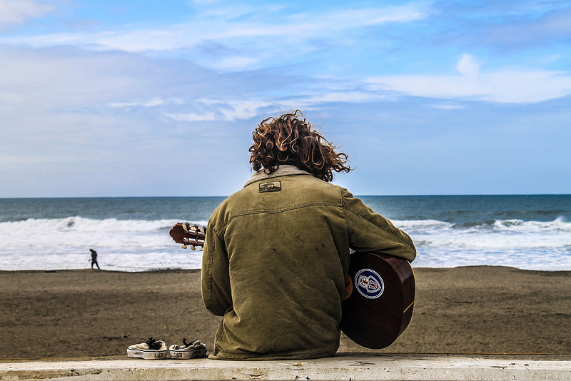 Man with his back to us plays the guitar on the beach