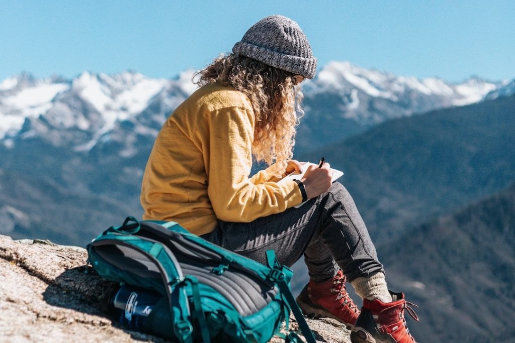 A girl sits on a rock with the mountains in the background writing in her journal