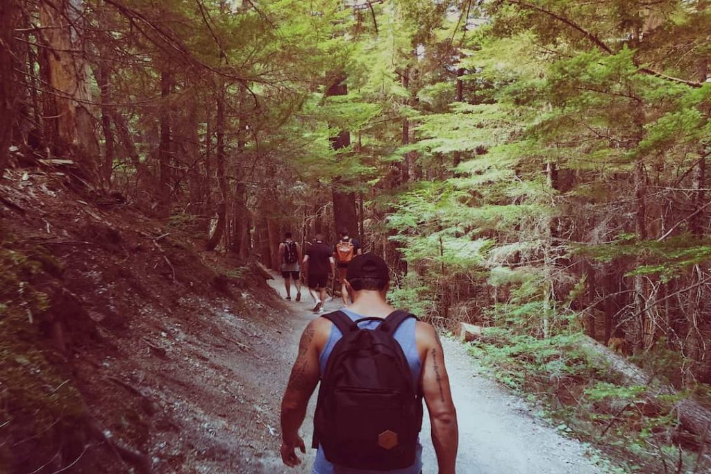 A man with tattoos wearing a backpack walks behind a row of people in a forest