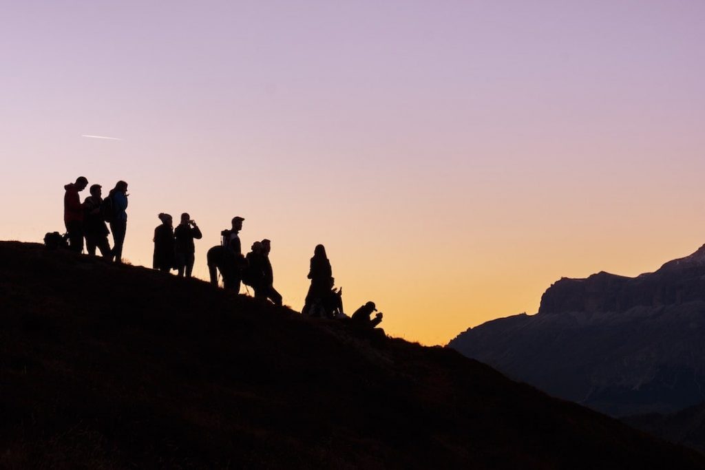 A group of people at sunrise or sunset standing on a large hill with a purple and orange sky in the background