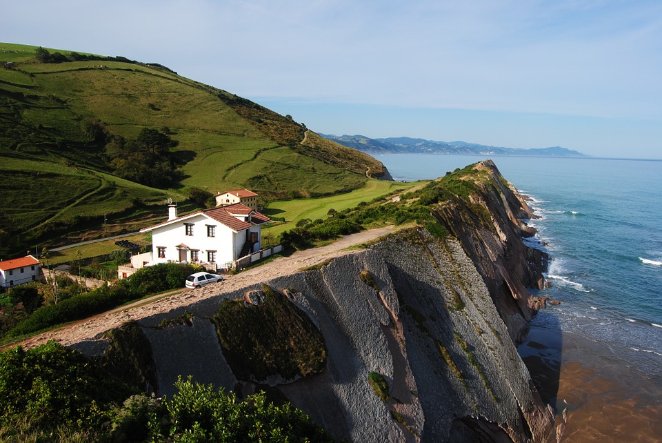 a car drives along the coast where a solitary white house sits on top of a cliff