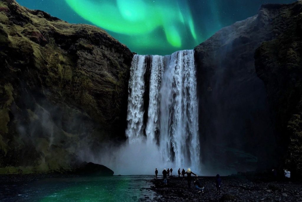 A group of people stand under Skógafoss in Iceland with the Northern Lights above