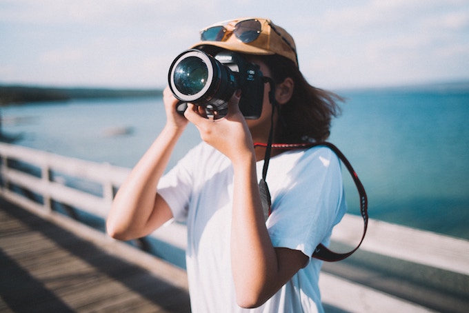 A person in a white shirt stands by a body of water taking a picture with a camera with a large lens