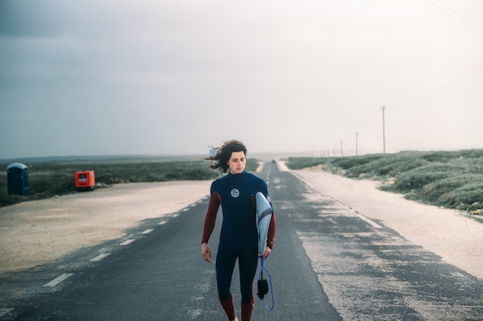 A person with long hair wearing a wetsuit walks down a sandy road holding a surfboard in Portugal