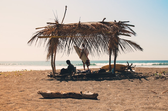 People holding surfboards sitting under a thatched roof structure on a beach in Costa Rica