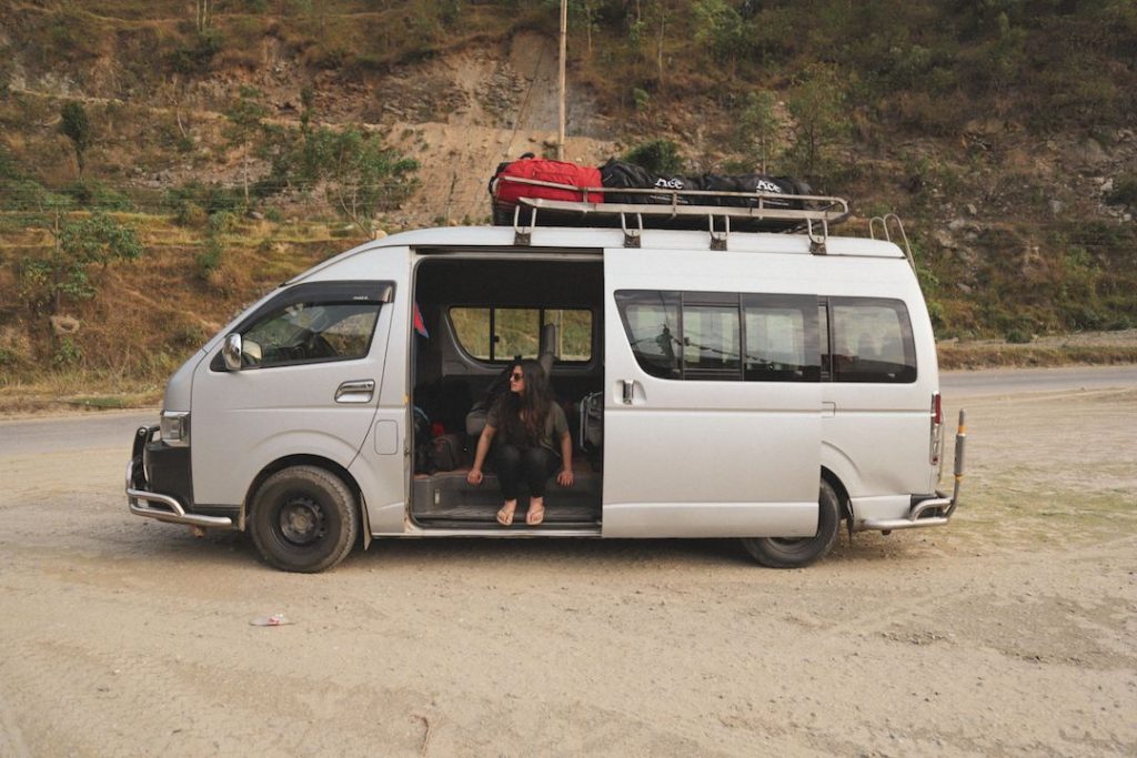 a girl sits in a white suzuki van which has red and black duffel bags on top of it