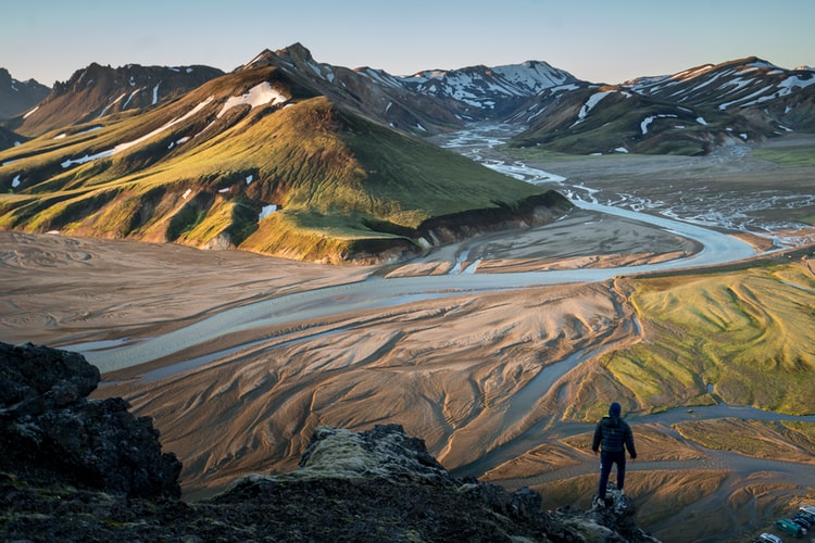 man in black jacket standing in front of a green valley with a river snaking through it