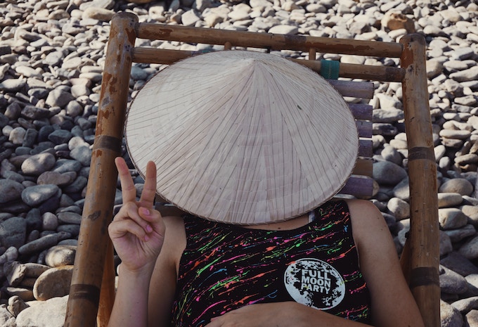 A man giving a peace sign under a hat on a beach in Thailand