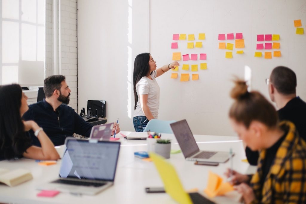 Woman standing at the front of a classroom pointing to post-it notes on the wall