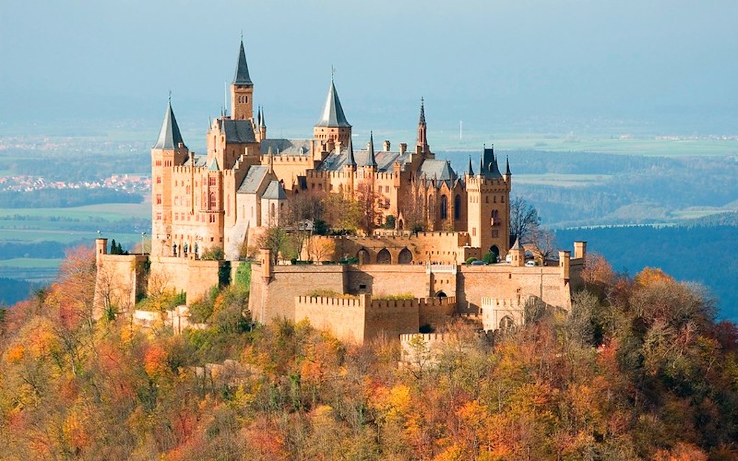 a castle on top of a hillside covered in trees with autumn colours of red, orange and brown