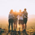 Group of friends with their arms around eachother and a sunset in front on them