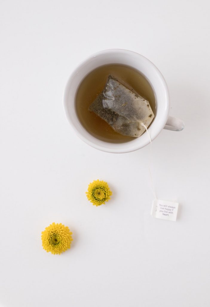 Cup of green tea with tag bag inside and two yellow flowers beside it