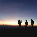 Three people with backpacks standing on a mountain at sunset