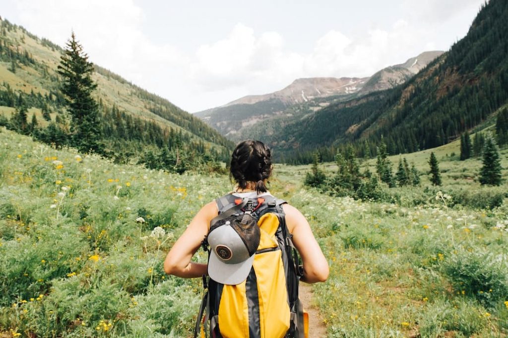 woman wearing yellow backpack standing in front of a hiking trail and mountain peaks