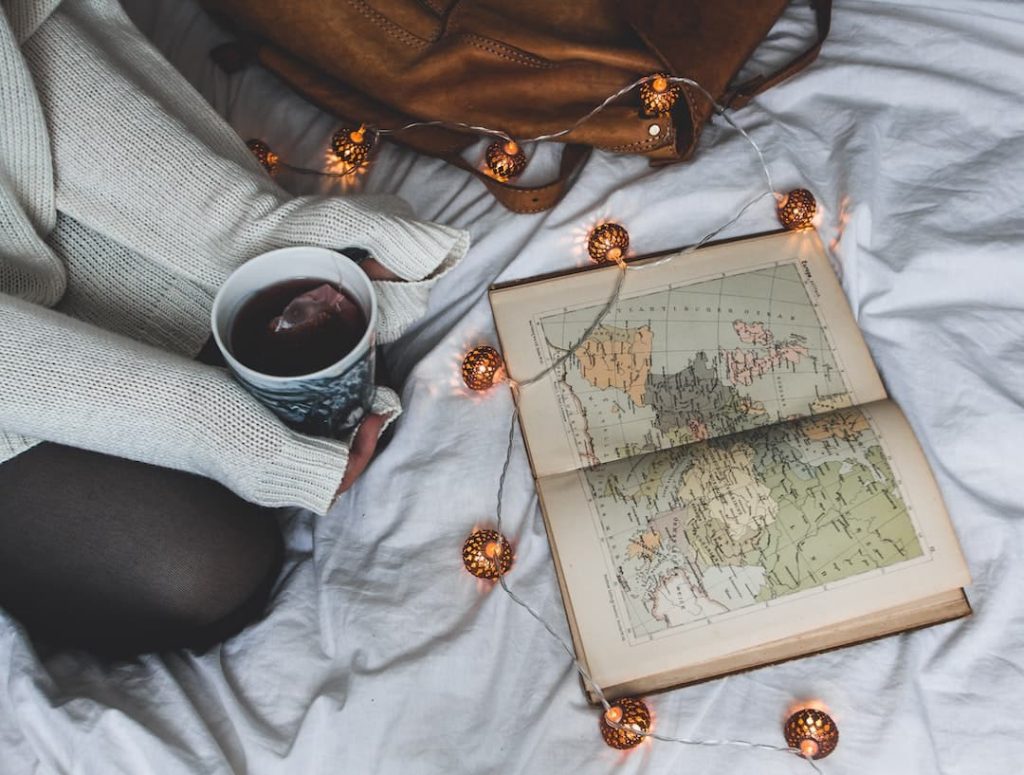 Person sitting on bed holding a cup of tea with an atlas open and fairy lights