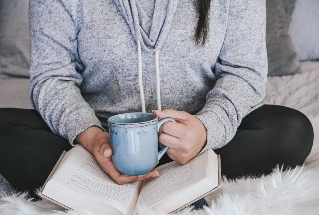 Person sitting on bed holding a cup of tea with a book in their lap