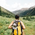 Woman with yellow backpack hiking through field with mountain peaks