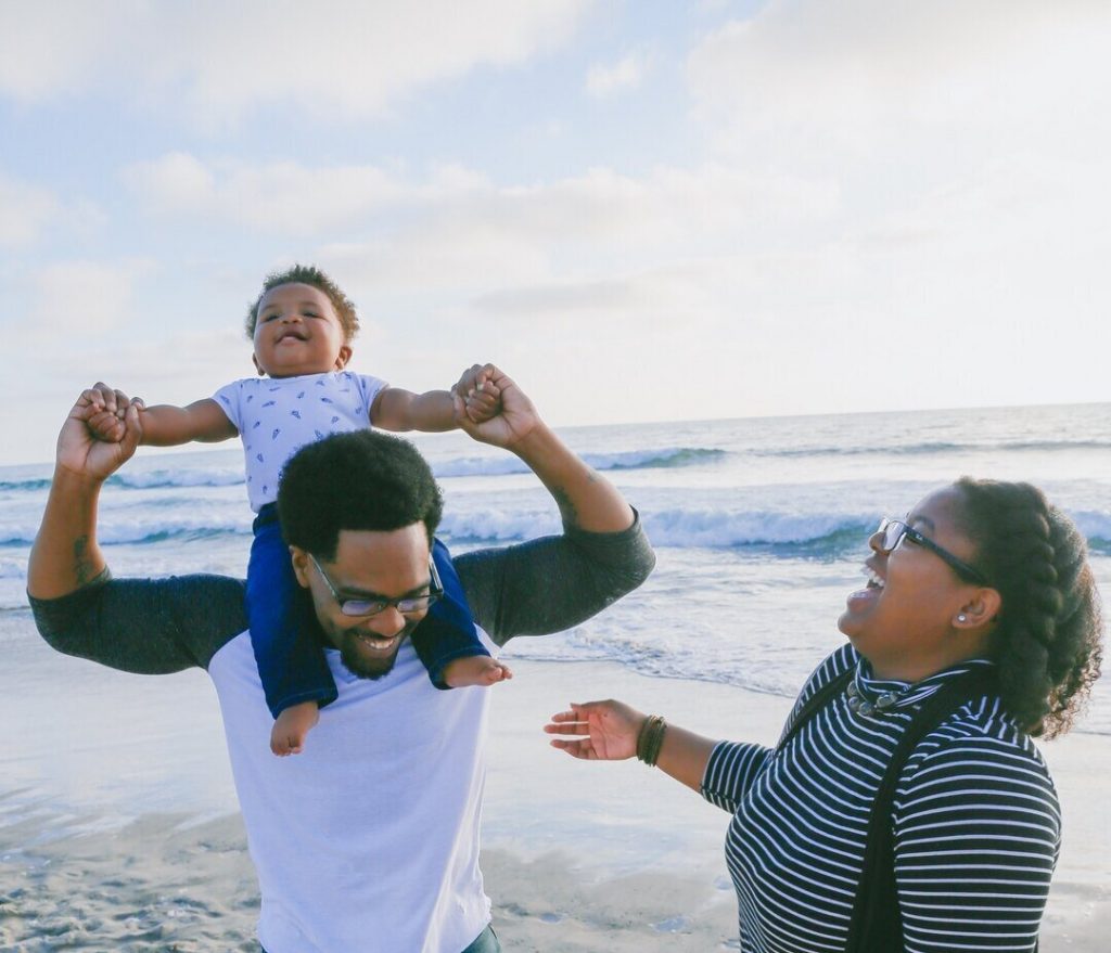 With the child sitting on his father's shoulders, the family of three stands happily on the beach and enjoys their private adventure