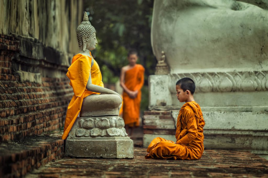 A young monk can be seen meditating in front of a Buddha statue  in a temple in Thailand.