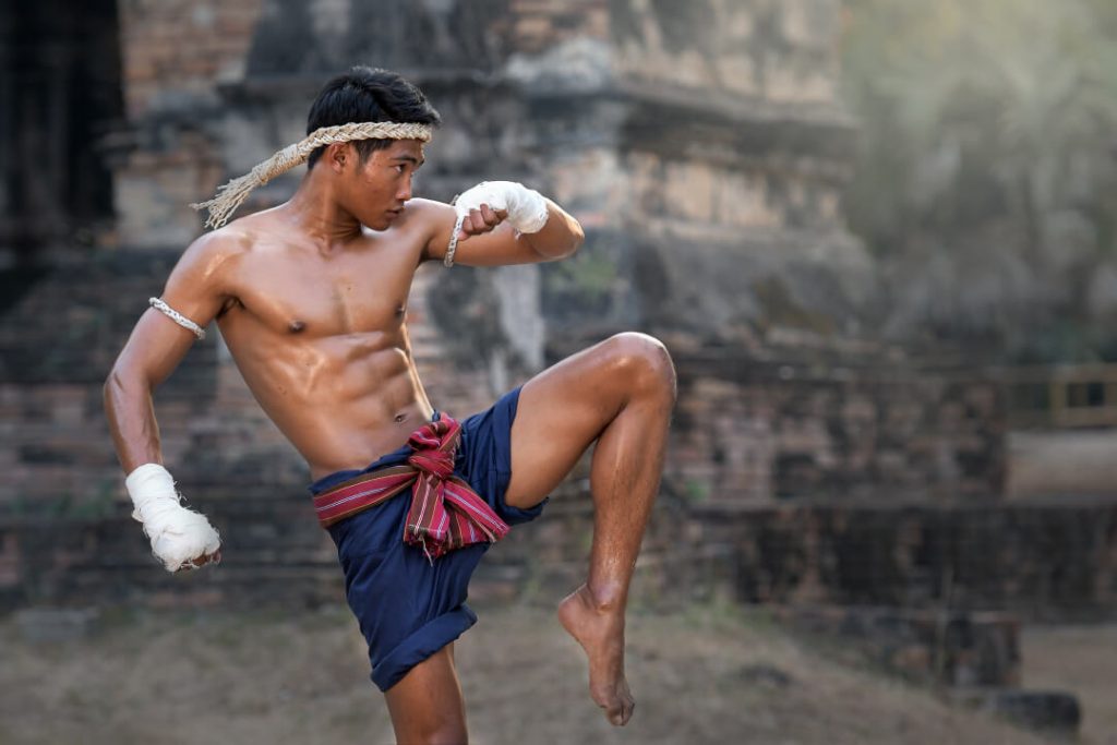 A young man stands with his left leg bent in a martial arts stance as he practices Muay Thai outside a temple in Thailand.