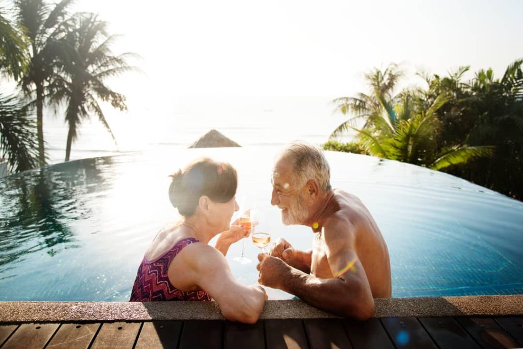 An older couple enjoys a glass of wine at a wellness retreat located in a beautiful setting with a thermal pool, palm trees, and the ocean in the background.