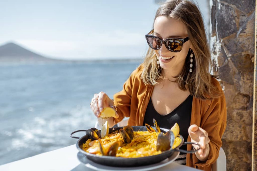 Explore the world through food like this woman who is eating paella, a traditional Spanish dish, while sitting in a seaside restaurant in Spain.