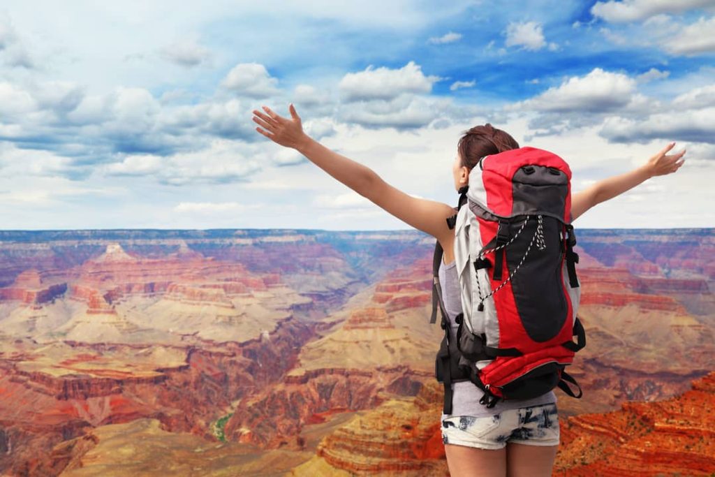 A happy woman with a backpack stands with arms raised as she looks out at her amazing view of the Grand Canyon.