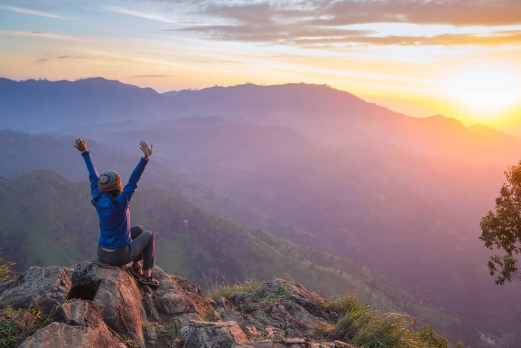 Happy woman sits with her back to us in nature (arms raised in celebration) as she stars out at a colorful sunset.